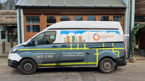 The side of a Wales and West Utilities van. The vehicle is parked on the side of a road, in front of businesses. The van has the gas utility logo printed on its side. It says 'your gas emergency and pipeline service'.