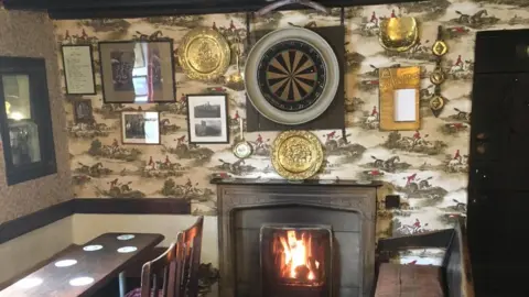 Glenys Crampton There is a fireplace with a fire in the centre of a room with a table and chairs to the left-hand side and a bench to the right-hand side. There is patterned wallpaper with horses and riders on the wall in the background. Hanging on the wall are several pictures and a dartboard.