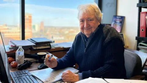 Jim Simpson sat in an office. He is a desk and holding a pen. He is wearing a blue jumper and has a keyboard, papers, an apple and a bottle of water nearby. He is smiling and a scene in Birmingham can be seen behind him.