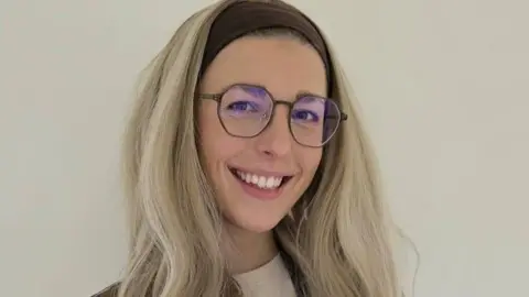 Wolverhampton Literature Festival A woman with glasses wearing a long blonde wing and smiling. She is holding a book.