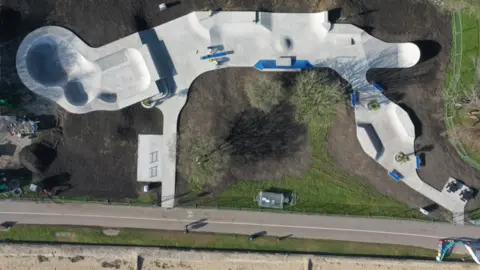 Aerial view of the skatepark, which is a light coloured concrete snaking through grass and trees beside the coastal path and beach