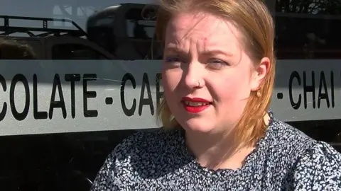 Andrew Sinclair/BBC Shona Hoey, wearing a black and white patterned dress or top, looks slightly off camera. She has red lipstick and auburn hair brushed behind her ears. Behind her is the window of a cafe.