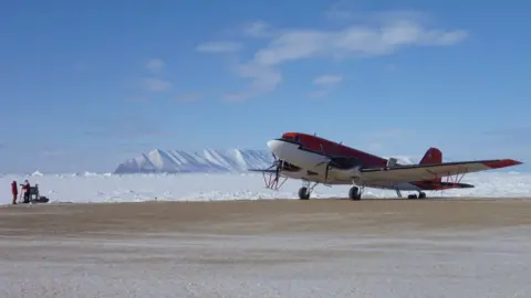 Tom Richter A red aeroplane stationary on a landing strip. Ice field and mountains visible in the background.