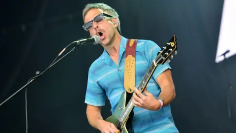  Irwin Sparkes of The Hoosiers performs on stage at Penn Fest on July 23, 2022 in Penn Street, Buckinghamshire. He is wearing sunglasses, a blue short sleeve shirt and holding a guitar as he sings into a microphone.