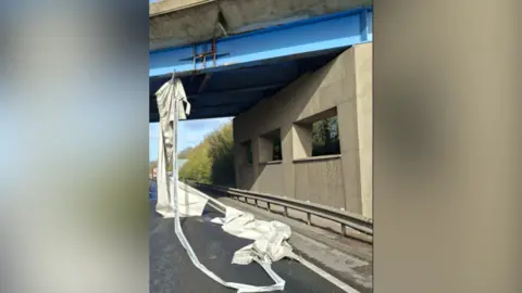 National Highways East A large piece of white material which has been torn off the side of a lorry hanging down from an overbridge on the A14. 