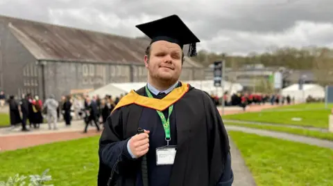A university graduate dressed in black gown and mortar board, with a yellow hood decoration. He has a sunflower lanyard around his neck and is holding a black walking aid. He stands on grass, with white marquees, other students and a large building out of vision behind him.