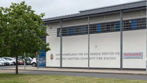 BBC The Northumberland Fire and Rescue service headquarters building. It is a white two-storey building with the words "Northumberland Fire and Rescue HQ" on the side and the red-and-yellow crest of the service. There is a grassy area at the front and a tree on the left of frame.