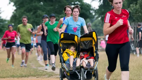 Reuters Runners dressed in t-shirts and shorts and running trousers in various colours head towards the camera while running on short grass. One woman runner pushes two boys in a double buggy. 