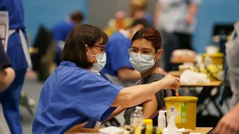 PA Media A person wearing a blue facemask sat next to a nurse in a facemask. The nurse is holding a needle and is about to administer a vaccine.