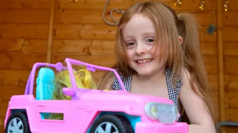 A little girl sits at a table with a toy car. She is smiling at the camera.