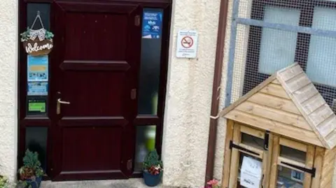 Helen Williams Markham playgroup entrance, a brown door is visible to the left and a wooden animal hutch with items like postcards is to the right. 