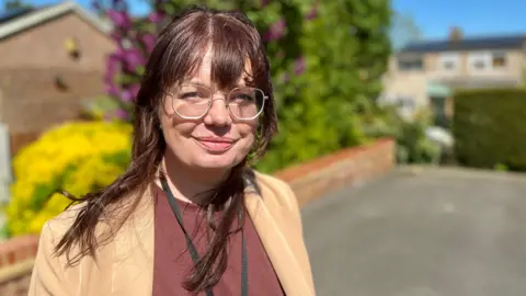 Claire Colvill, a social work apprentice, looks at the camera outside a client's home in Hertfordshire. She is wearing a light brown suit jacket with a maroon smart T-shirt underneath it. She has a black lanyard around her neck and is wearing metal framed glasses. She has brown hair with a fringe that has a red tone. She is standing in front of a driveway with a brick wall and hedges along the wall.