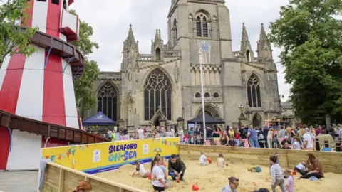 Children playing in a pop-up beach in Sleaford's market place. A cathedral and a red and white helter-skelter are in the background. People are sat on deckhairs.