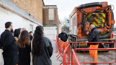 A group of students with their backs to camera listening to a trainer who is gesturing towards a construction vehicle