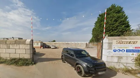 A black car is leaving a car park on an industrial estate next to a sign saying Towens. Concrete block walls surround the site.