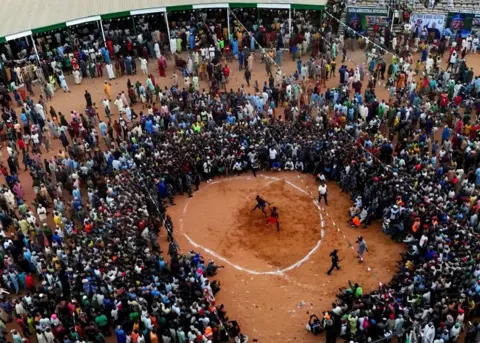 Reuters A drone view shows people gathering to watch Dambe fighters compete in the ancient Nigerian combat sport . A white circle drawn on the ground can be seen and the two fighters are inside.