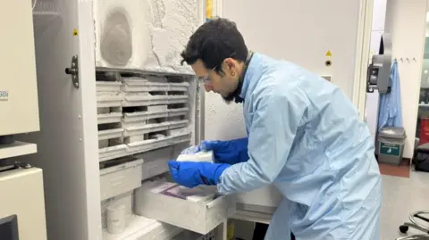 A man in a pale blue lab coat, goggles and thick blue temperature resistant gloves is reaching into a freezer in a lab. It contains several drawers and containers all covered with a thick layer of frost and ice. 