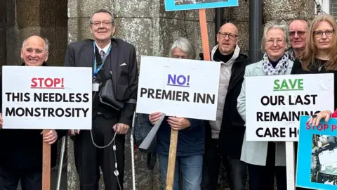 About 15 people stood outside an old looking hotel. Many of them are holding placards with phrase such as 'No Premier Inn' and 'Stop the needless monstrosity'. There is a bench to the bottom right of the photo and a flower bed to the left. 