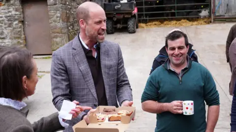The Prince of Wales wearing a grey suit jacket stood in a courtyard of a farm. He is holding a cardboard box of baked goods. A lady in a grey fleece coat laughs to his left and a man in a green quarter zip jumper holding a cup of tea smiles and laughs to his right.