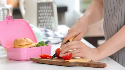 Getty Images Mother cutting tomato for school lunch at table in kitchen (stock photo)