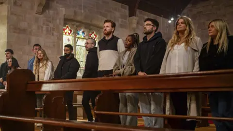  BBC/Studio Lambert/Llara Plaza The cast of the Traitors lined up in a church. L-R Harry, Charlotte, Andrew, Jaz, Zack, Paul, Jasmine, Ross, Charlie and Evie. The cast stand in pews inside a stone church with stained glass windows visible behind them