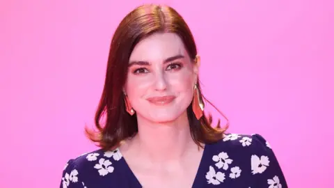 Getty Images Laurie Nunn, pictured in 2021. Laurie is a white woman in her 30s with dark brown hair reaching her shoulders. She has a side parting and wears her hair loose. She has brown eyes and smiles as she looks at the camera. She is wearing a dark blue V-necked dress with a white floral print and large dangling gold and orange earrings. She is pictured in front of a bubble-gum pink background.