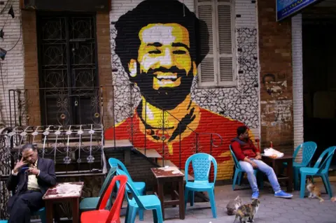 Reuters A mural of Egypt and Liverpool football star Mohamed Salah is seen as people sit outside a street coffee shop downtown in the capital of Cairo, Egypt January 16, 2018.