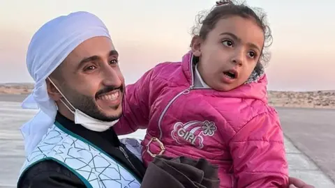 An aid worker is carrying a small child across the tarmac