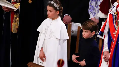 Yui Mok Princess Charlotte is wearing a white cape with embroidery and silver headpiece. Prince Louis is wearing a dark blue leather tunic