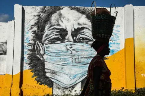 AFP A Tanzanian woman carries a basket on her head as she walks in front of a mural depicting a masked person.