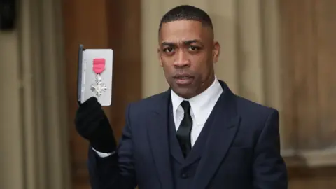 Getty Images Wiley, also known as Richard Cowie, poses with his medal after being appointed an MBE in 2018