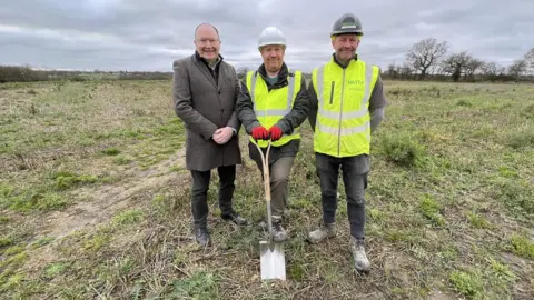 Stuart Woodward/BBC Three people stood in a field with a spade, marking the start of construction of the Maldon crematorium.