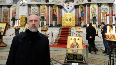 Ioann Kurmoyarov Father Ioann stands in front of Orthodox Church icons.