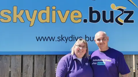 Sarah Luxton Sarah Luxton and her brother Wayne Ewing standing in front of a skydive banner with purple shirts on that say 'rebuilding lives after stroke'