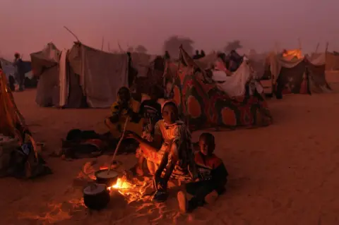 Dan Kitwood/Getty Images Newly arrived Sudanese refugees cook food over fires outside their makeshift shelters at twilight close to a relocation camp on April 24, 2024 near Adre, Chad.