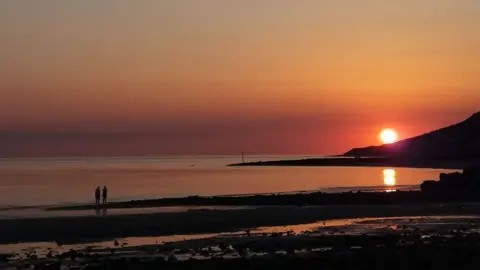 Win Watson Silhouette of couple in distance on shoreline with sun setting and orange sky over coast