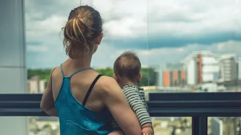 Getty Images Mother and baby looking out of their apartment at city beyond