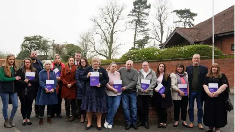 PA Media Chelsey Campbell, Carley McKee, Colin Griffiths, Fiona Carr, Charlotte Cheshire, Rhiannon Davies, Richard Stanton, Kayleigh Griffiths, Donna Ockenden, Nicky Lauder, David Boylett, Hayley Matthews, Steph Hotchkiss, Julie Rawlings, Neil Rawlings and Sonia Leigh stand with the final Ockenden report at The Mercure Shrewsbury Albrighton Hotel, Shropshire.