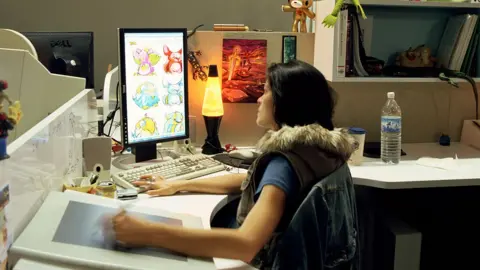 Getty Images An artist using a drawing tablet on a computer, sat in an office. Her desk has a lava lamp as well as a screen on which six Neopets are visible.