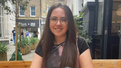 INSTAGRAM/SOPHIACAREY Sophia, wearing glasses, is smiling at the camera. She is sat down surrounded by shops, with plants and trees in the background