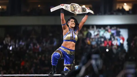 WWE Bayley holds her winner's belt aloft after winning WWE Women's Champion at WrestleMania. Bayley is a 34-year-old white woman with long brown hair. She wears a blue and gold crop top with matching blue trousers and black boots.