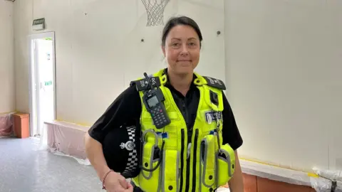 Officer Gemma Rutter smiling at the camera. She has dark hair and is wearing a hi-vis police vest with a radio attached to it. She is standing in the redecorated scout hall