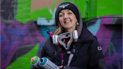 Hannah Constance Hannah Constance stands in front of an artwork while she holds spray paint cans