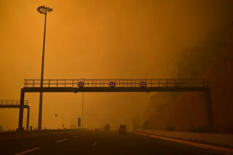 Getty Images A road block is set up amid smoke as a wildfire burns in Kineta, near Athens, on July 23, 2018