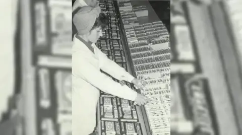 Private Collection A woman puts together a selection box of confectionery, wearing a pristine white overall