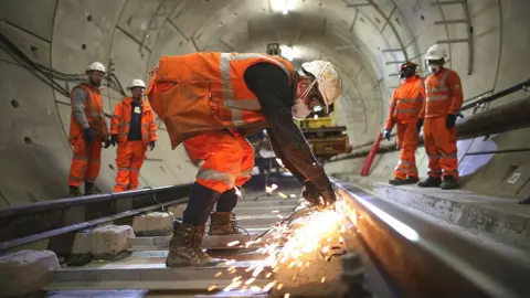 Getty Images Construction workers wearing white hard hats and orange hi-viz protective clothing work inside a tunnel for the Elizabeth Line underground line on 16 November, 2016.