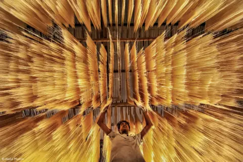 Abdul Momin Man reaching up to rice noodles that are hanging above him