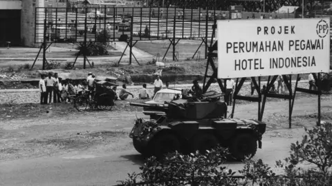 Getty Images Young people stood outside an hotel in Indonesia with a military armoured vehicle on stand by