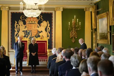 PA Media Alex Maskey delivers a message of condolence to King Charles and Camilla inside the Throne Room at Hillsborough Castle as guests watch the proceedings