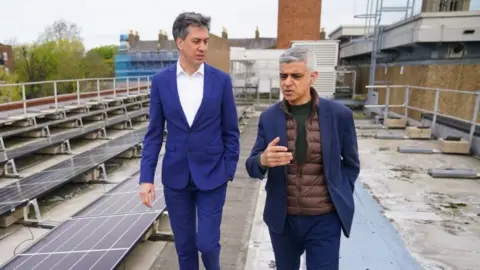 PA Media Ed Miliband listening to Sadiq Khan talking as they walk on the roof of a building with solar panels